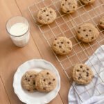 a glass of milk beside a wire cooling rack filled with chocolate chip cookies and a hand towel next to a white plate of cookies