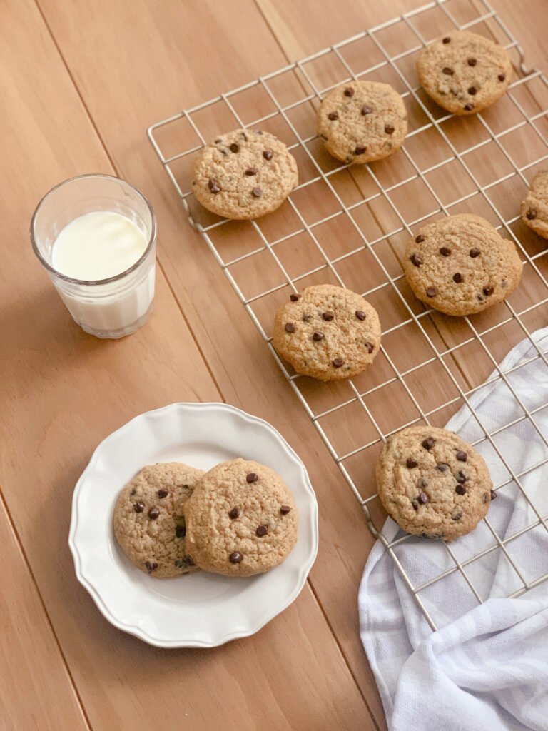 a glass of milk beside a wire cooling rack filled with chocolate chip cookies and a hand towel next to a white plate of cookies