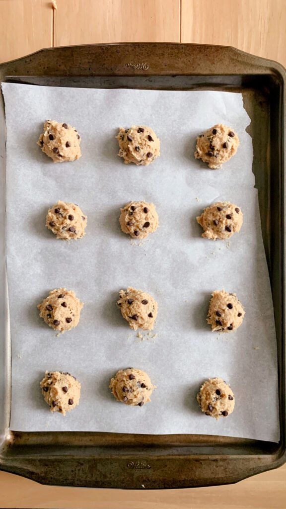 12 fresh milled chocolate chip cookie dough balls evenly spaced on a layer of parchment paper in a baking tray