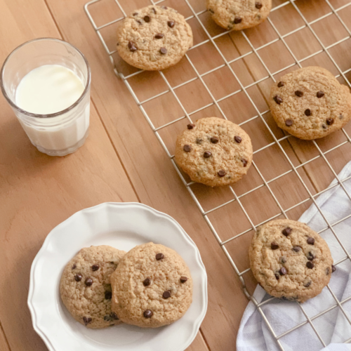 a glass of milk beside a wire cooling rack filled with chocolate chip cookies and a hand towel next to a white plate of cookies