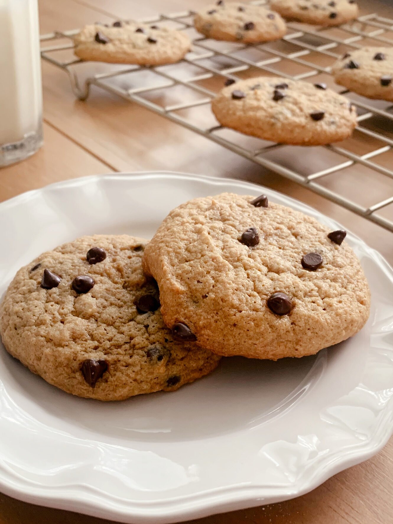two fresh milled chocolate chip cookies on a white plate near a cooling rack filled with cookies