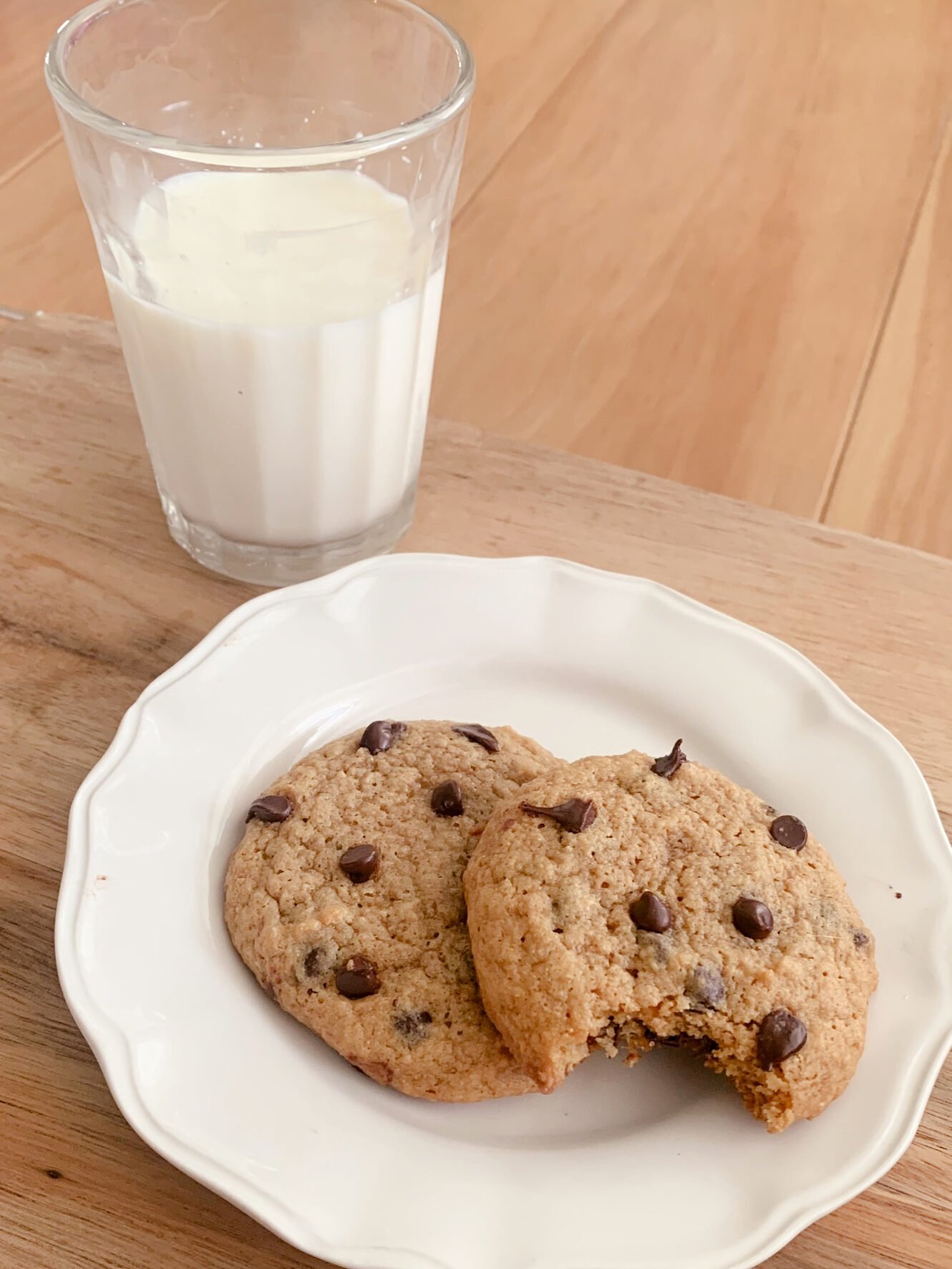 A glass of milk next to two fresh milled chocolate chip cookies sitting on plate, one has a bite out of it.