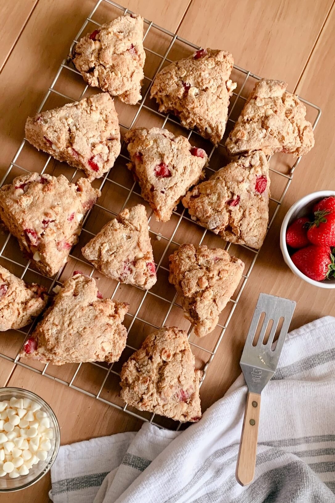 fresh milled white chocolate strawberry scones on a wire rack next to a bowl of strawberries, bowl of white chocolate chips, metal server, and tea towel on a wooden table.