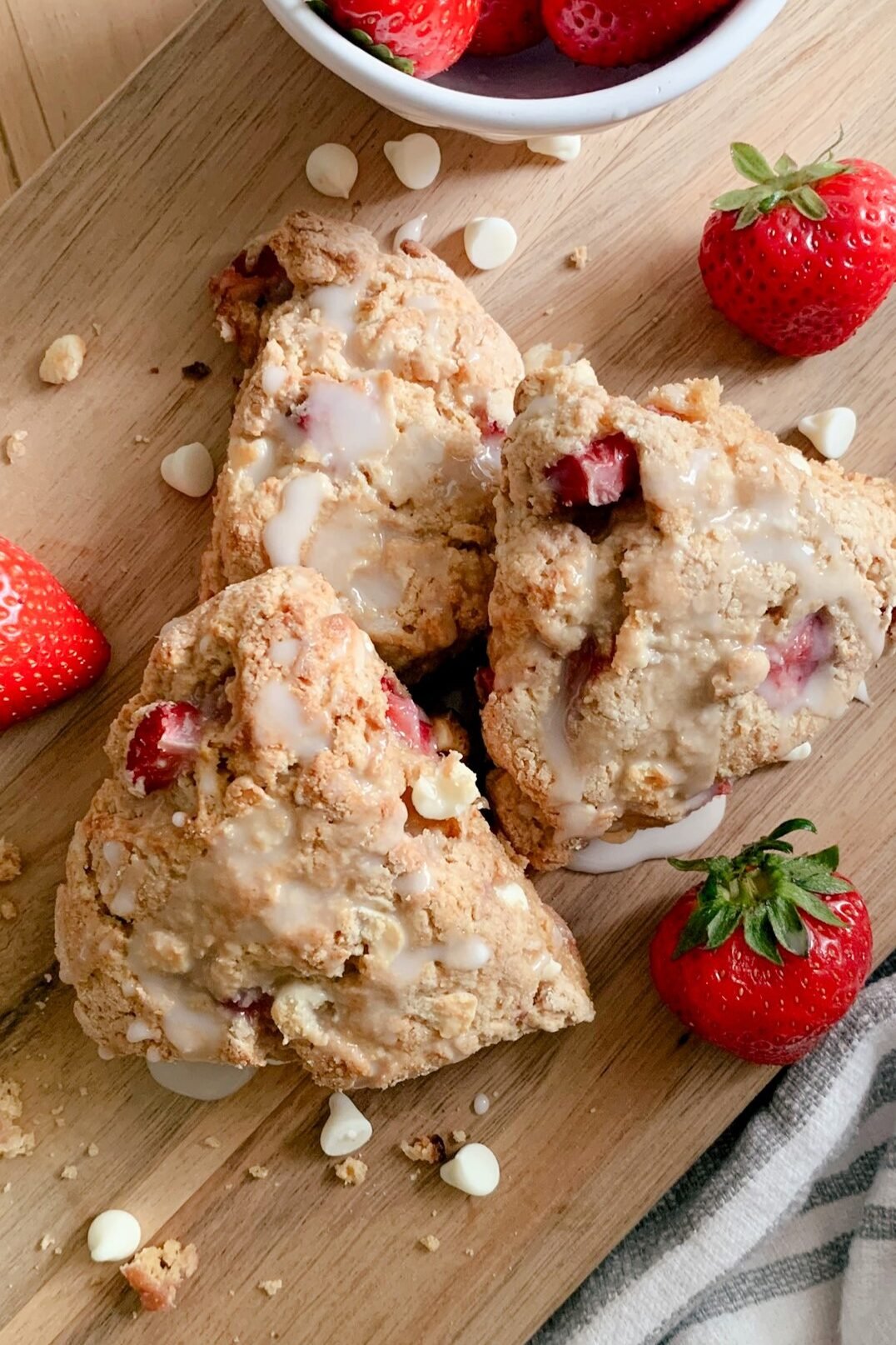 Fresh milled white chocolate strawberry scones layered on a wooden cutting board with strawberries, white chocolate chips, crumbs next to a tea towel