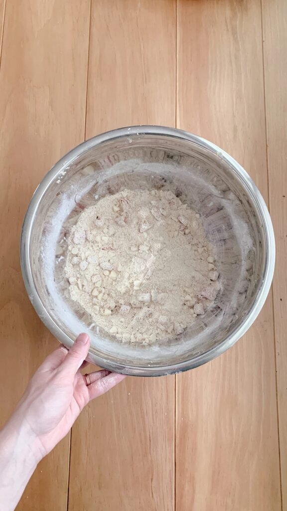 a hand holding a stainless steel bowl of ingredients for fresh milled white chocolate strawberry scones on a wooden table