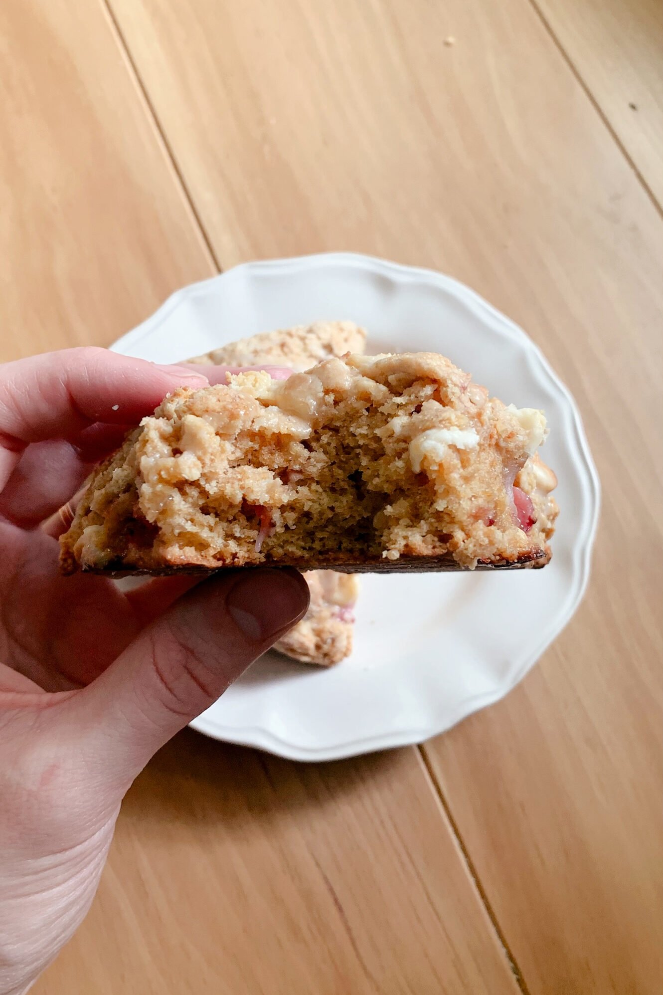 a hand holding a bitten fresh milled white chocolate strawberry scone in front of a white plate on a wooden table