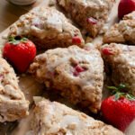 fresh milled white chocolate strawberry scones drizzled with icing on a wooden cutting board with strawberries and a bowl of white chocolate chips on a wooden table