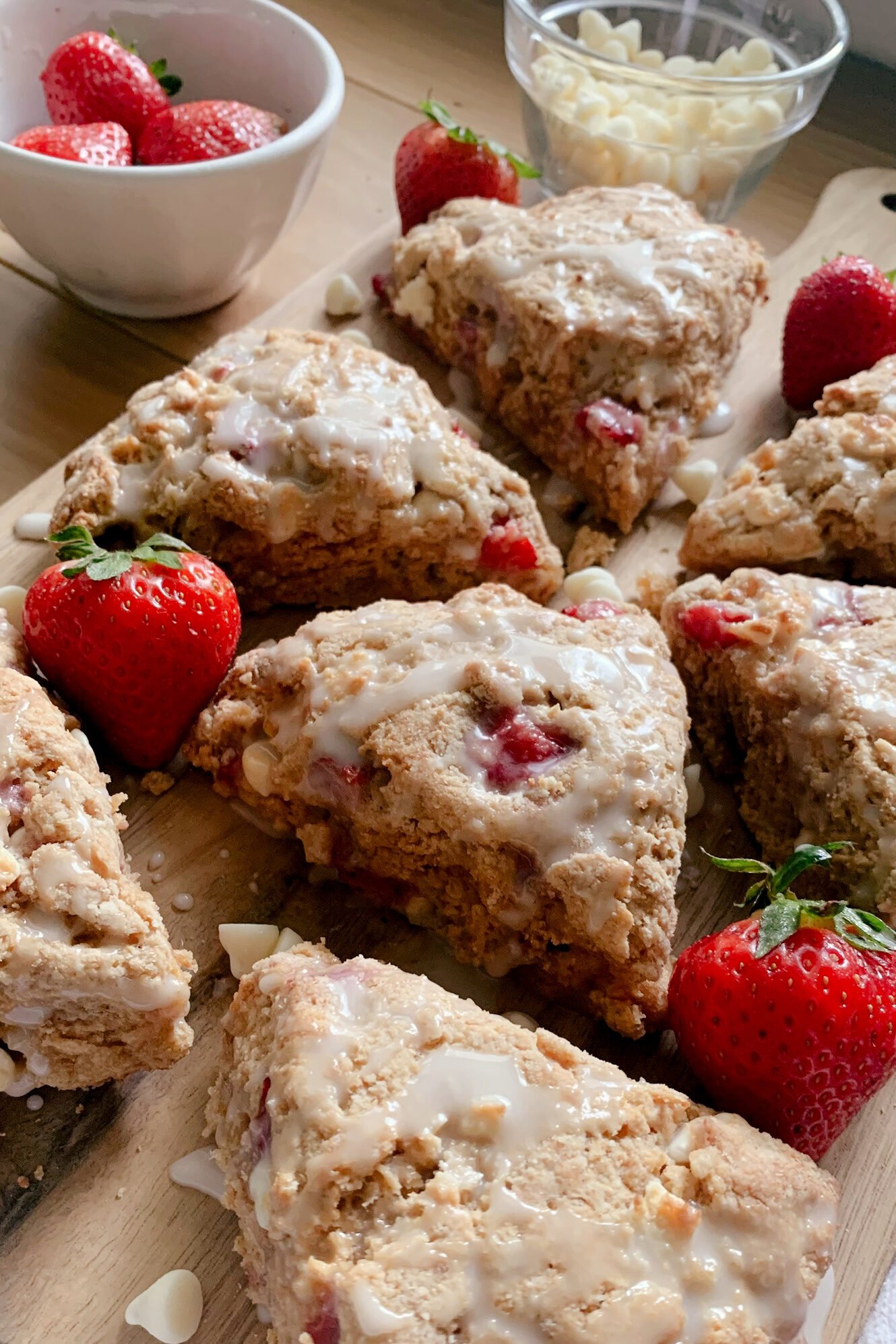 fresh milled white chocolate strawberry scones drizzled with icing on a wooden cutting board with strawberries and a bowl of white chocolate chips on a wooden table