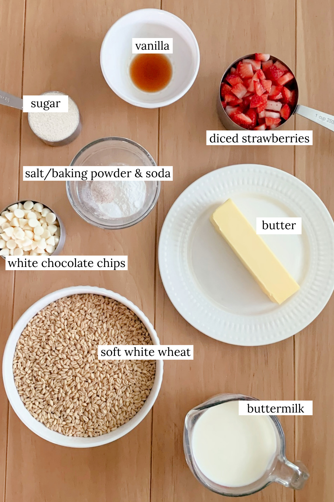 Ingredients for fresh milled white chocolate strawberry scones in various measuring cups, plates, and bowls on a wooden table